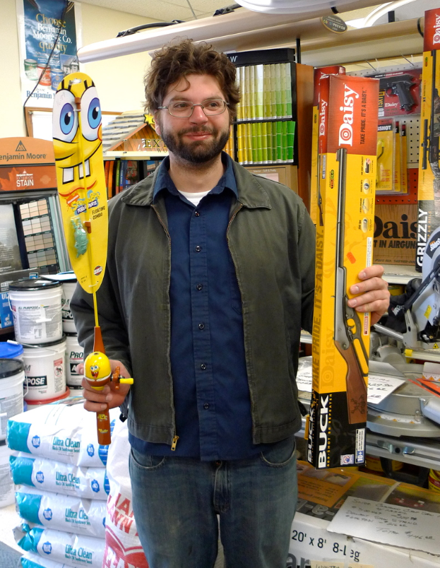 Eine Person mit Brille hält eine Box mit einem Gewehrbild und Text sowie einem weiteren Gegenstand in den Händen, zusammen mit verschiedenen Gegenständen wie Eimern und Paketen und einem Plakat an der Wand im Hintergrund.