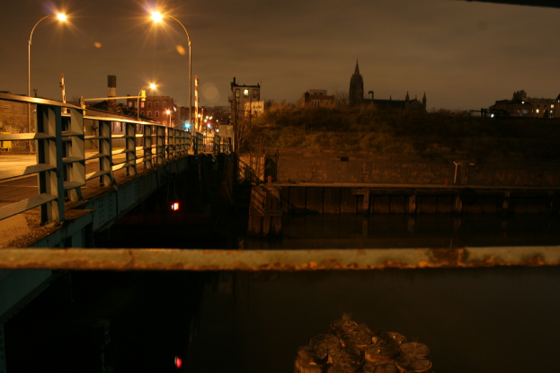 Eine Brücke überspannt einen Fluss mit zahlreichen Straßenlaternen darauf und Gebäude sind im Hintergrund zu sehen.