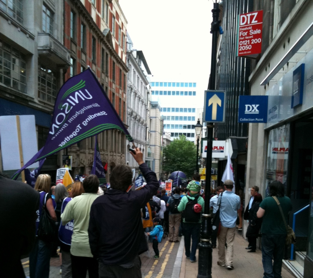 Eine große Gruppe von Menschen auf einer Straße, einige halten Fahnen und Schilder, mit einem Schildständer auf einem Pfahl, Gebäuden mit Fenstern an den Seiten und einem Baum, Laternenmast, Gebäude und Himmel im Hintergrund.