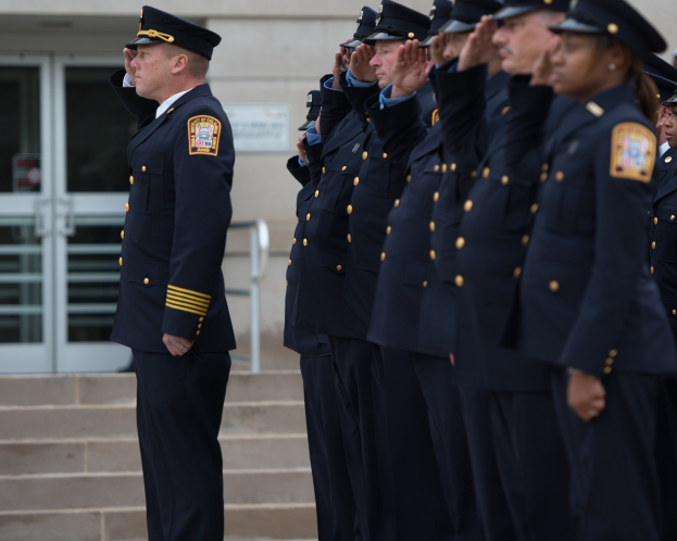Eine Gruppe von Polizisten in Uniform und Mützen, die in Formation salutieren vor einem Gebäude mit Glastüren und Treppen.