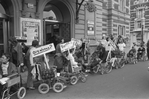 Eine Gruppe von Menschen im Rollstuhl mit Schildern an der Straße, im Hintergrund Gebäude und ein Laternenmast auf der rechten Seite, in Schwarz-Weiß.