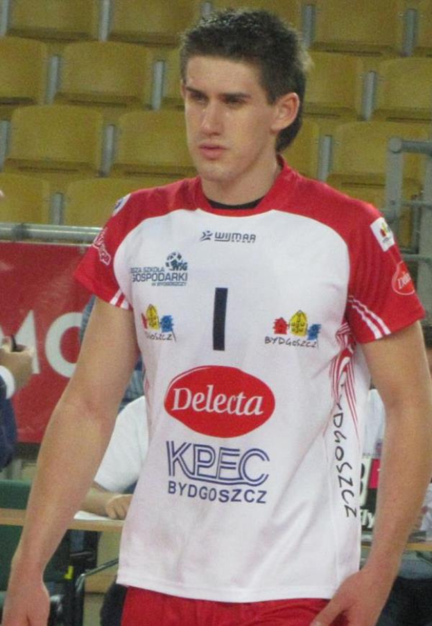 Ein Mann in einem roten und weißen Handballtrikot steht auf einem Feld, sieht entschlossen aus, mit Zuschauern und einem roten Banner mit der Aufschrift "Krasnoyarsk vs Bydgoszcz" im Hintergrund.