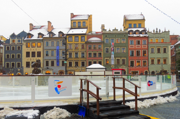 Eislaufbahn in einem städtischen Platz mit schneebedecktem Boden, umgeben von Gebäuden, Laternen, Bäumen und einem Himmel als Hintergrund.