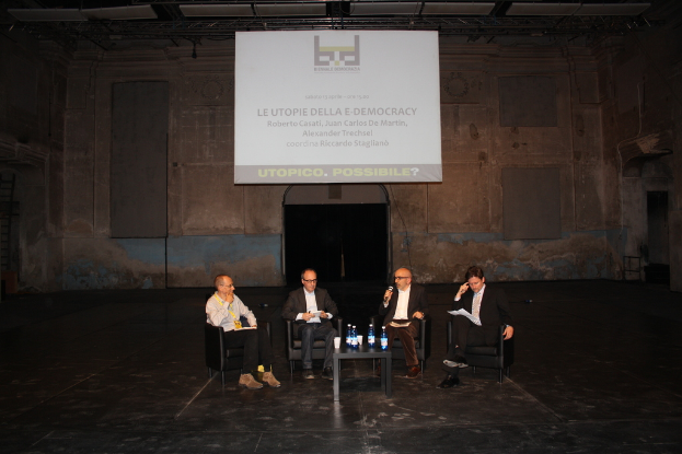 Vier Personen sitzen in Stühlen, eine spricht in ein Mikrofon, mit einem Tisch vor ihnen, der Wasserflaschen und Gläser enthält, und einem Projektor, der ein Bild hinter ihnen anzeigt.