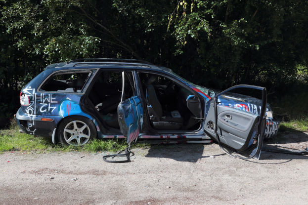 Ein verunfalltes Auto am Straßenrand mit Gras und Bäumen im Hintergrund.
