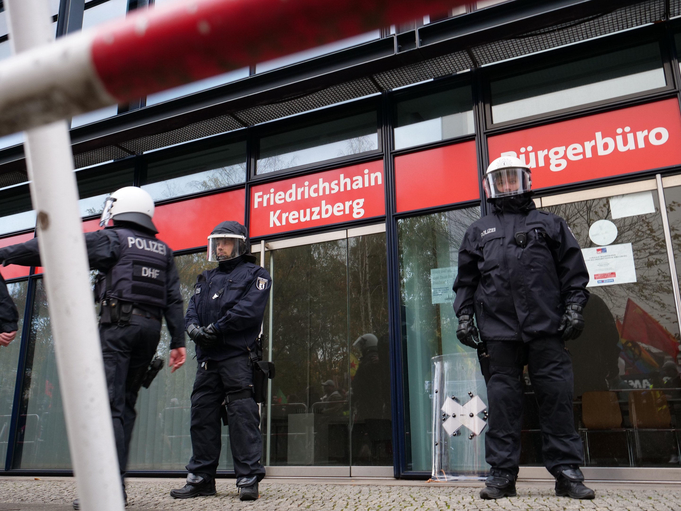 Polizisten in Helmen und Handschuhen vor einem Glaswandgebäude mit angeklebten Papieren und einem Pfahl links; Hintergrundtafeln zeigen "Friedrichshain Kreuzberg."