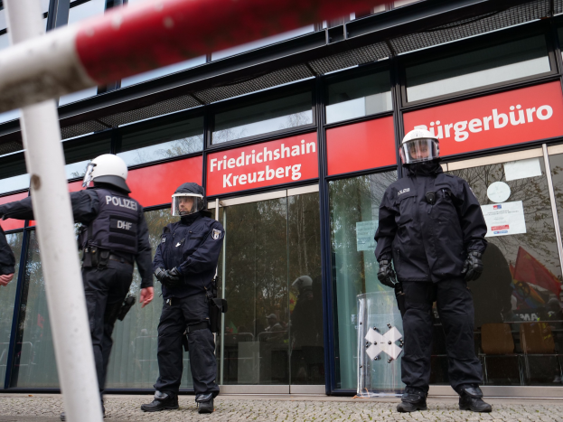 Polizisten in Helmen und Handschuhen vor einem Glaswandgebäude mit angeklebten Papieren und einem Pfahl links; Hintergrundtafeln zeigen "Friedrichshain Kreuzberg."