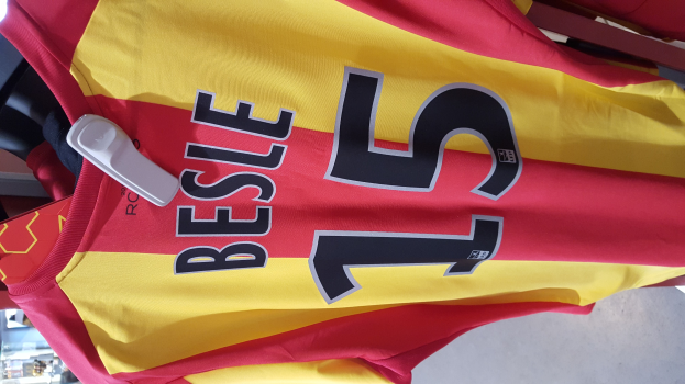 Rotes und gelbes Fußballtrikot mit aufgedrucktem Text und Zahlen, das an einer Ladengestell hängt, mit anderen Gegenständen und dem Boden im Hintergrund.