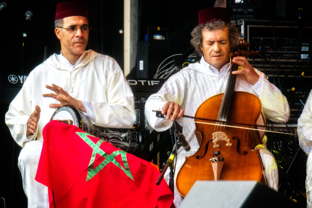Zwei Männer in Mützen und passenden Kostümen sitzen und spielen verschiedene Musikinstrumente, wobei einer eine Brille trägt und Musikgeräte hinter ihnen sichtbar sind.