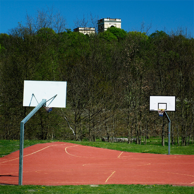 Ein Spielplatz mit Gras, zwei Basketballstangen, Bäumen, einem Gebäude im Hintergrund und einem klaren Himmel.