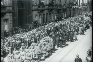 Schwarzes Foto einer Parade mit einer großen Menge Menschen, die eine Straße entlang gehen, einige halten Gewehre in den Händen, vor einem Gebäude mit einem Wasserzeichen in der rechten oberen Ecke.