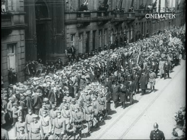 Schwarzes Foto einer Parade mit einer großen Menge Menschen, die eine Straße entlang gehen, einige halten Gewehre in den Händen, vor einem Gebäude mit einem Wasserzeichen in der rechten oberen Ecke.