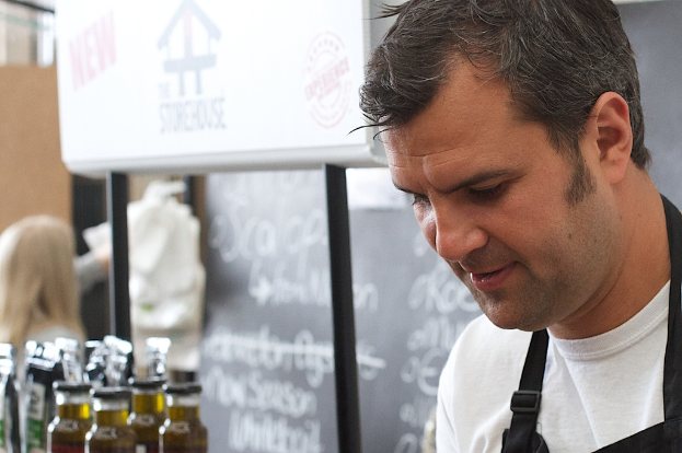 Ein Mann in einem weißen Hemd steht rechts im Bild vor einer Tafel, während eine Frau hinter Flaschen links steht und ein Schild in der Mitte positioniert ist.