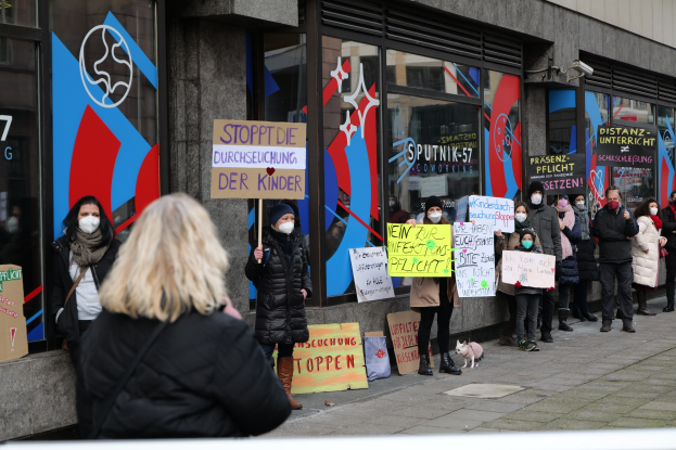 Maskierte Menschen mit Schildern und Plakaten vor einem Glasgebäude, mit Kameras und einem Hund, scheinen gegen eine deutsche Regierungspolitik zu protestieren.