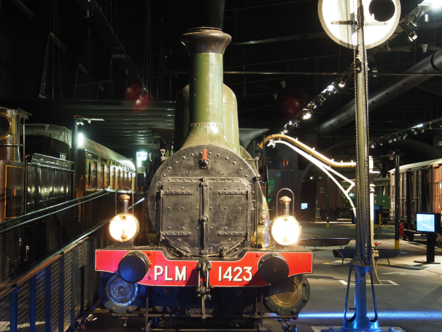 Eine Dampflokomotive in einem Museum ausgestellt, von einem Metallzaun umgeben, mit Besuchern, die sie beobachten.