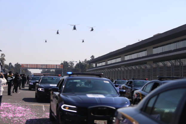 Eine Gruppe von Polizeiwagen vor einem Gebäude geparkt, mit Menschen auf der linken Seite der Straße, Bäumen, Pfählen, einer Brücke und einem klaren blauen Himmel im Hintergrund und vier Hubschraubern, die oben im Bild fliegen.
