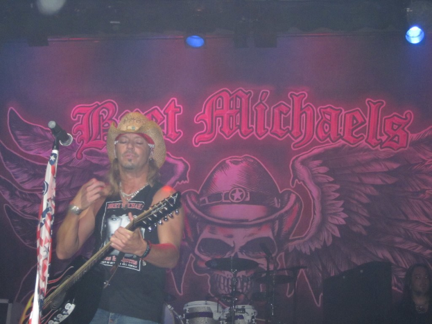 Eine Konzertszene mit einem Gitarristen links neben einem Mikrofon, einer Person in der unteren rechten Ecke, einem Banner im Hintergrund und Drogen in der Mitte unten, mit Beleuchtung von oben.