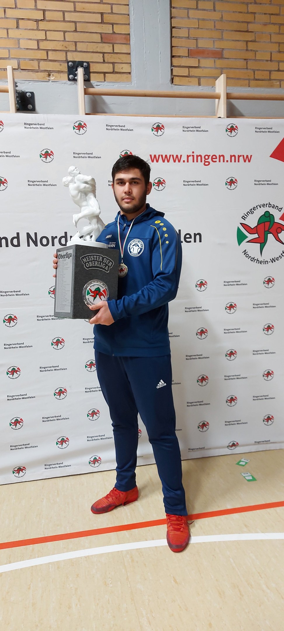 Ein Mann in einem Anzug hält einen Pokal, während er eine Medaille trägt und vor einer bannergeschmückten Wand steht.