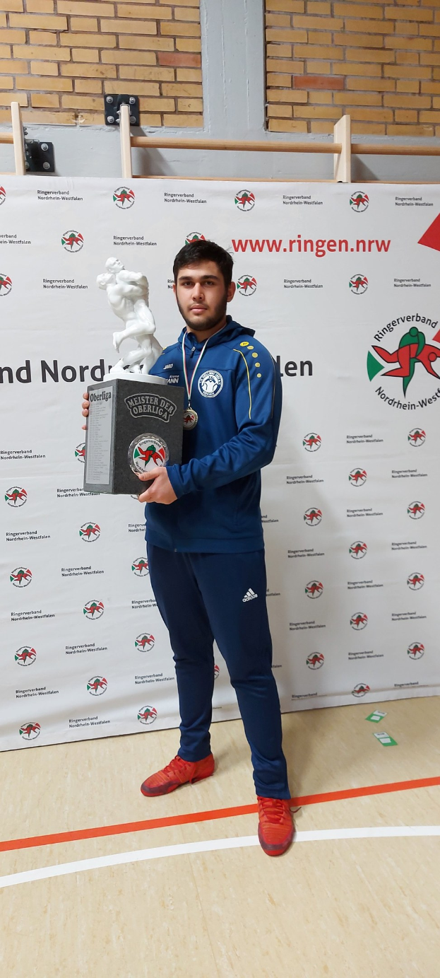 Ein Mann in einem Anzug hält einen Pokal, während er eine Medaille trägt und vor einer bannergeschmückten Wand steht.