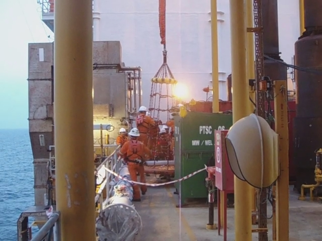 Ein Schiff mit Masten, Kisten und Geländern, das Menschen in Helmen zeigt, vor einem Wasserhintergrund.