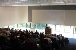 Ein Seminarraum in einer Universität mit vielen Menschen, die aufmerksam einer Präsentation zugewandt sind, die vorne angezeigt wird, wo eine Person an einem großen Tisch die Inhalte erklärt, mit Glasfenstern im Hintergrund, die Bäume und Gebäude zeigen.