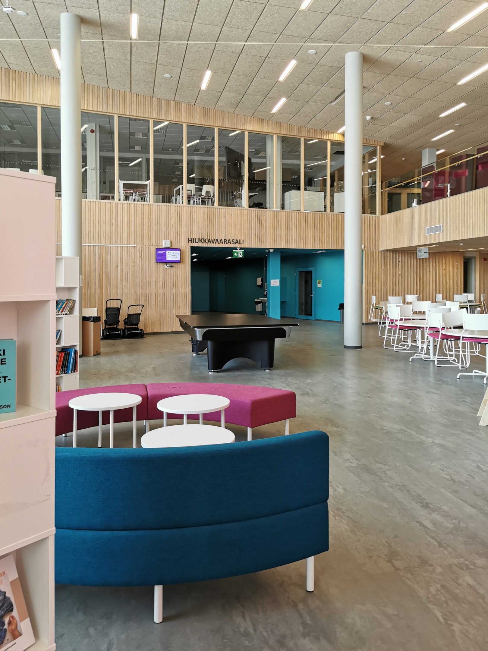 Großer Raum mit mehreren Tischen, Stühlen, Sofas, Hockern, Bücherregalen, Säulen, Deckenleuchten und einem Wandfernseher, der an ein Schulbibliotheks-Interieur erinnert.