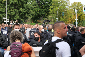 Eine große Gruppe von Menschen steht vor einer Menschenmenge aus Polizeibeamten, einige tragen Masken und Rucksäcke, andere halten Kameras, mit Laternenmasten, Verkehrssignalen, Schildern, Bäumen und einem klaren blauen Himmel im Hintergrund.