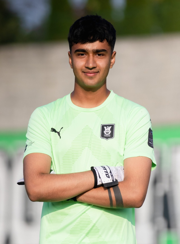 Ein junger Mann in einem grünen Hemd steht auf einem Fußballfeld mit verschränkten Armen, lachend, mit einem unscharfen Hintergrund.