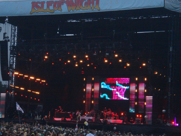 Eine Bühne mit Lichtern, ein Bildschirm und Musiker, die Instrumente spielen, mit einem Banner oben, einer Menge unten und Fahnen mit Stangen und Ballons drumherum.