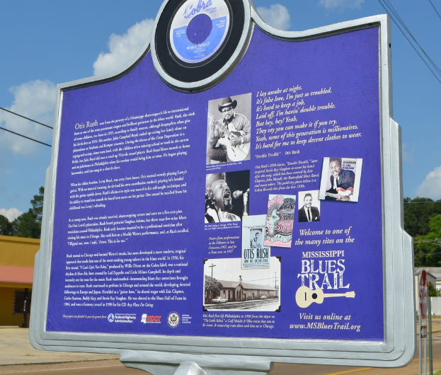 Ein Infobrett, Gebäude, ein Schuppen, Bäume, Stromkabel und ein bewölkter Himmel.