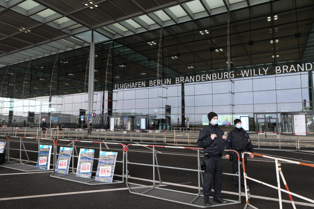 Zwei Polizisten in schwarzer Uniform vor einem Glasgebäude mit Text, mit Absperrungen und Schildern im Vordergrund.