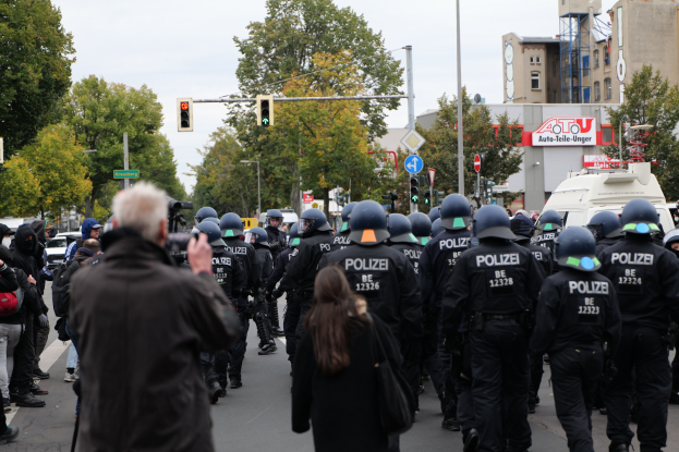 Eine Gruppe von Menschen geht auf einer Straße mit Polizeibeamten, einige tragen Helme und tragen Taschen oder Kameras, vor einer Kulisse von Fahrzeugen, Verkehrsampeln, Schildern, Bäumen, Gebäuden und einem klaren blauen Himmel.