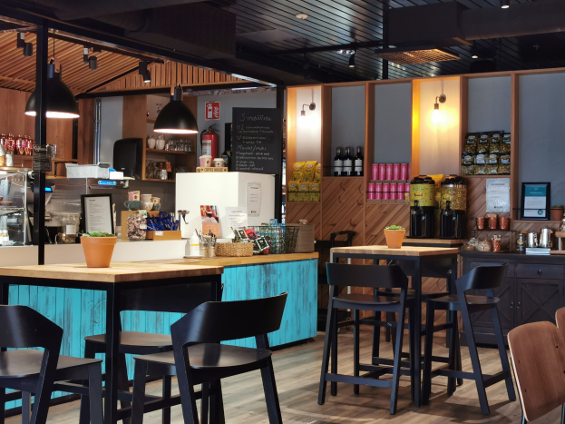 Innenraum eines Restaurants mit Holztischen und -stühlen, einem Topf mit einer Pflanze auf einem Tisch, einer Schiefertafel, einem Kühlschrank, einem Feuerlöscher, Regalen mit Flaschen, Schränken mit Gegenständen, einem gerahmten Foto an der Wand, Deckenleuchten und Lampen.