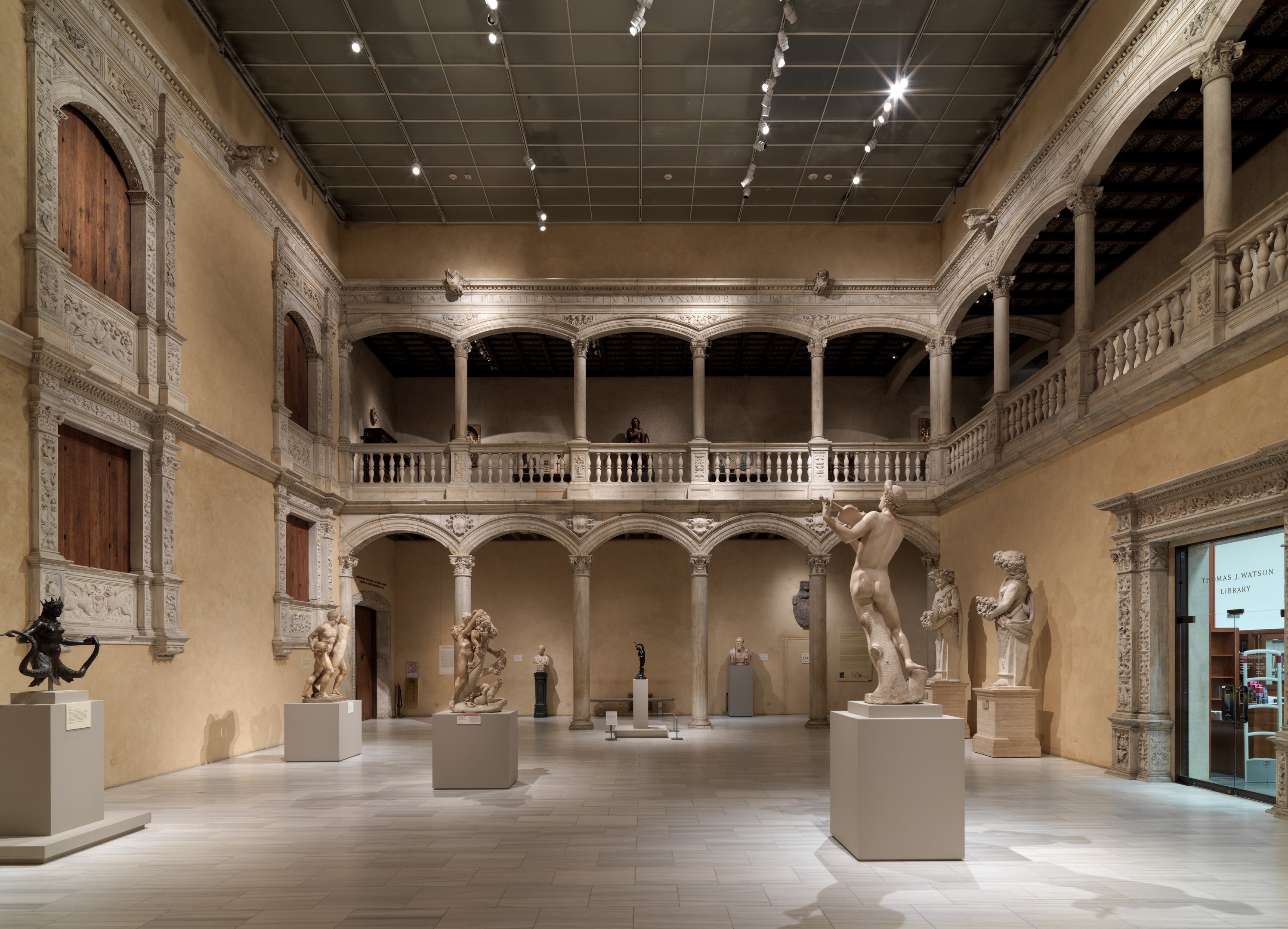 Großer Raum in einem Museum mit zahlreichen Statuen und Skulpturen auf Sockeln, architektonischen Elementen wie Säulen und Geländern, Deckenleuchten und einigen Besuchern.