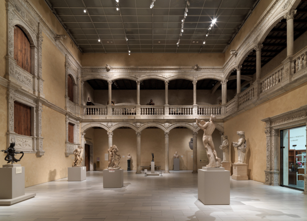 Großer Raum in einem Museum mit zahlreichen Statuen und Skulpturen auf Sockeln, architektonischen Elementen wie Säulen und Geländern, Deckenleuchten und einigen Besuchern.