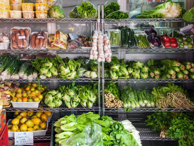 Ein Gang in einem Supermarkt mit frischem Obst und Gemüse, verpackten Artikeln in Plastikfolie, Kartons, ein Textschild und ein Glasfenster im Hintergrund.
