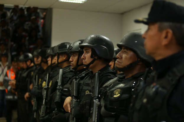 Polizeibeamte in Helmen und Uniformen mit Waffen, die in einer Reihe mit einer Wand und Deckenbeleuchtung im Hintergrund stehen.