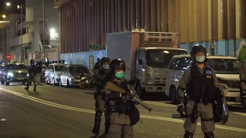 Polizeibeamte in Helmen, Handschuhen und Gesichtsmasken stehen auf einer Straße bei Nacht, einige halten Waffen, mit Fahrzeugen, Laternenmasten, Schildern und Gebäuden im Hintergrund.