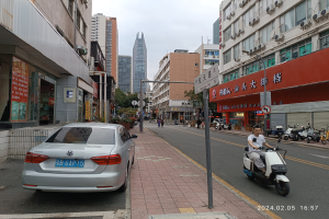 Ein Mann fährt mit einem Roller eine städtische Straße entlang, gesäumt von hohen Gebäuden, mit Menschen auf den Gehwegen und Fahrzeugen auf der Straße, unter einem klaren blauen Himmel mit Bäumen im Hintergrund.