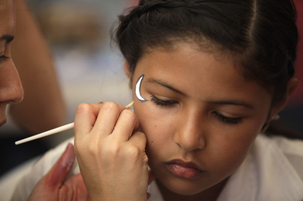 Ein Make-up-Künstler trägt Make-up mit einem Pinsel auf das Gesicht eines jungen Mädchens auf, wobei beide Personen vor einem unscharfen Hintergrund im Fokus stehen.