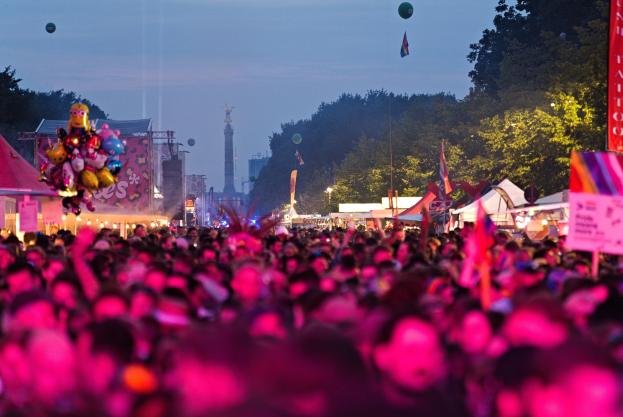 Große Menschenmenge bei einem nächtlichen Musikfestival mit farbigen Lichtern, Ballons, Zelten, Bannern, Flaggen, Bäumen, einem Turm und einem sternenklaren Himmel.