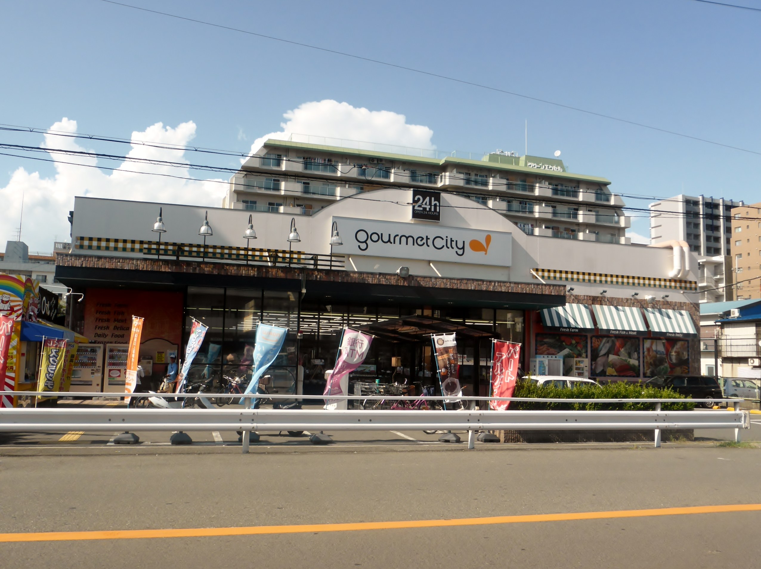 Eine belebte Stadtstraße mit Gebäuden, Straßenlaternen, Werbetafeln, Fahrzeugen, Absperrpollern, Büschen, Bäumen und einem bewölkten Himmel, in dessen Mitte ein Gourmetrestaurant mit verschiedenen Speisen zu sehen ist.