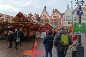 Eine Gruppe von Menschen geht eine Kopfsteinpflasterstraße neben einem Weihnachtsmarkt in Nürnberg, Deutschland, entlang mit Laternenmasten, Texttafeln und Gebäuden mit Fenstern im Hintergrund unter einem bewölkten Himmel.