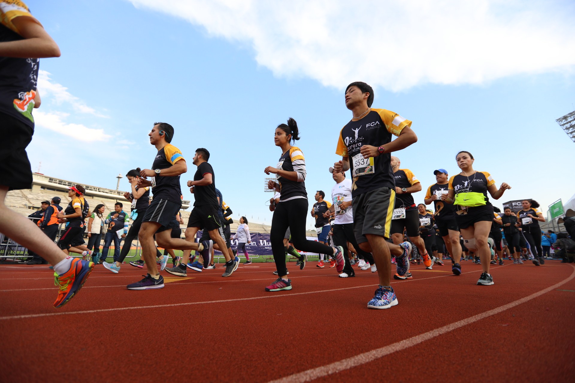 Eine Gruppe von Menschen, die auf einer Laufbahn laufen, mit einer Zuschauermenge auf der rechten Seite, vor Gebäuden, Laternenmasten, Bannern und einem klaren blauen Himmel.