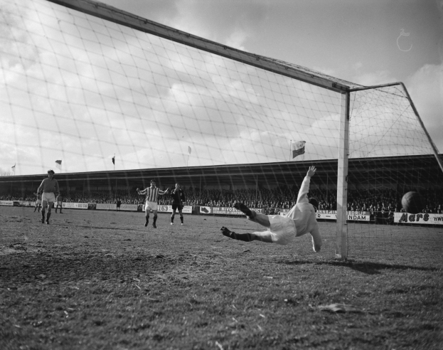 Ein Schwarz-Weiß-Foto eines Fußballspielers, der vor einem Tor einen Ball hält, umgeben von Spielern auf dem Boden und Zuschauern, Bannern und Fahnen im Hintergrund bei einem bewölkten Himmel.