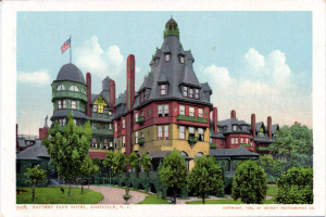 Eine alte Postkarte zeigt das Battery Park Hotel in Asheville, North Carolina, ein großes Gebäude mit einer Fahne oben drauf, umgeben von Bäumen, Pflanzen und Gras, mit einem Weg im Vordergrund und Text unten.