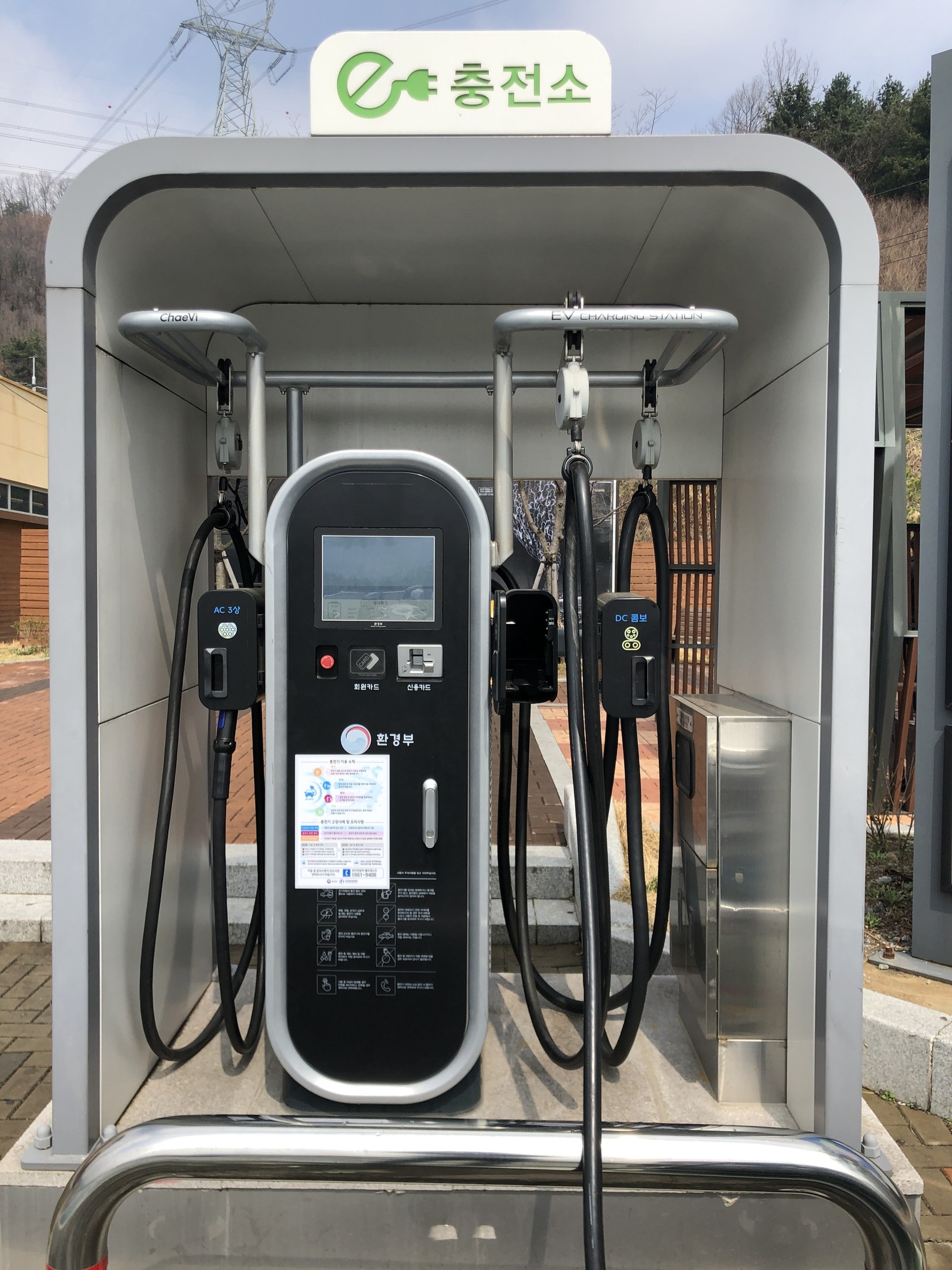 Elektrofahrzeug-Ladestation in Seoul, Südkorea, mit einer zentralen Lademaschine, verbundenen Kabeln und einer Texttafel, vor Buildings, Bäumen und einem Himmel-Hintergrund.