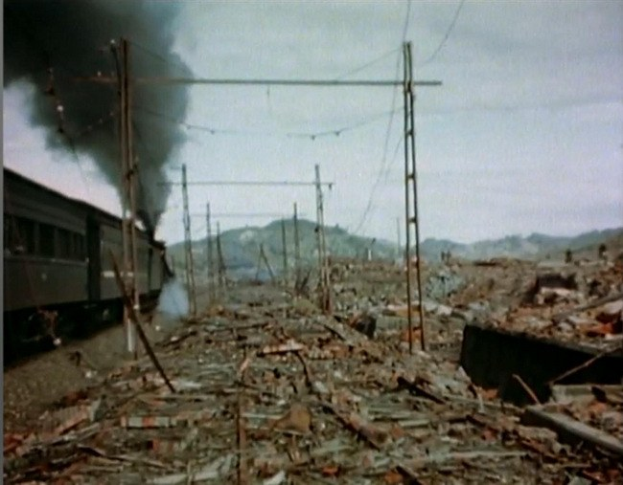Ein Zug fährt durch ein beschädigtes Gebiet mit Rauch, der aus ihm aufsteigt, umgeben von Strommasten und -drähten, mit Hügeln und einem klaren Himmel im Hintergrund und verstreuten Trümmern auf dem Boden.