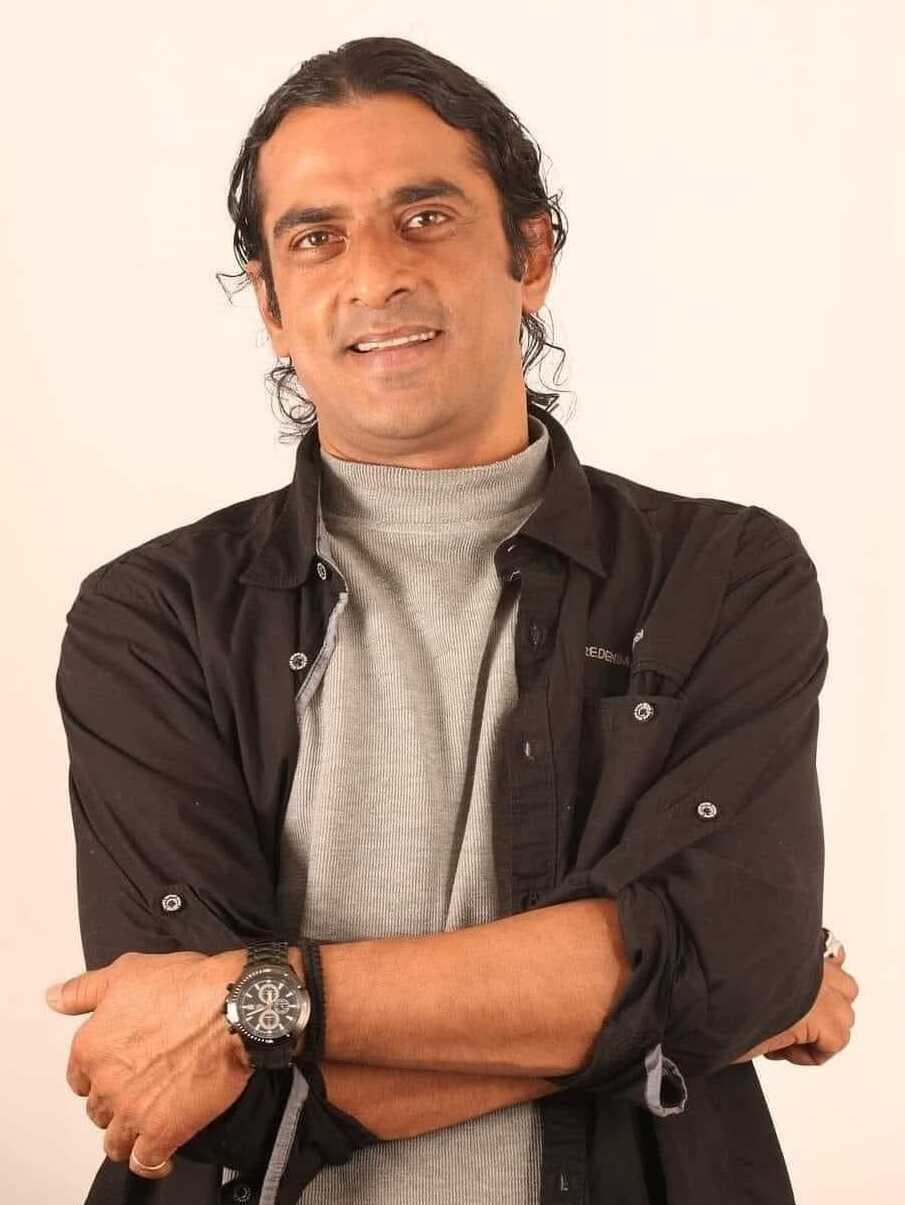 Ein Mann in einem schwarzen und aschgrauen Kleid mit verschränkten Armen vor einem weißen Hintergrund, der eine Uhr trägt und nachdenklich erscheint.