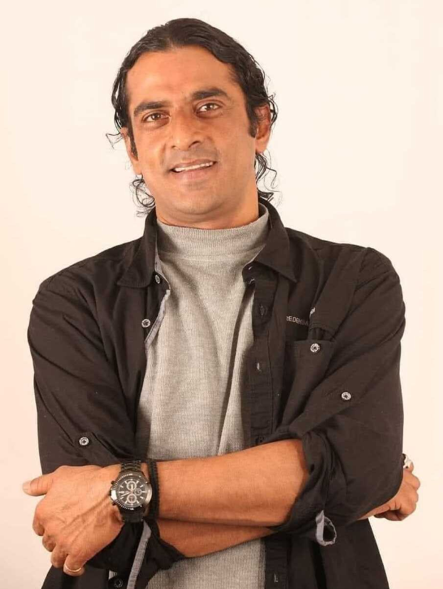 Ein Mann in einem schwarzen und aschgrauen Kleid mit verschränkten Armen vor einem weißen Hintergrund, der eine Uhr trägt und nachdenklich erscheint.